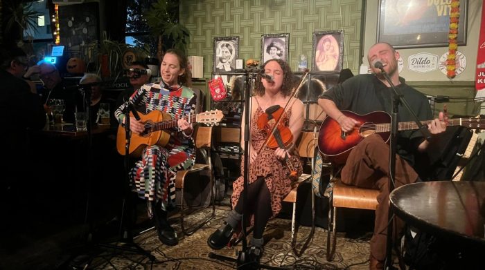 A trio of musicians performing live in a cozy venue. One woman plays guitar and sings, wearing a colorful patterned outfit. A second woman plays the violin and sings, dressed in a patterned dress. The third musician, a man, accompanies them on guitar. The background features framed photographs and a casual bar setting.